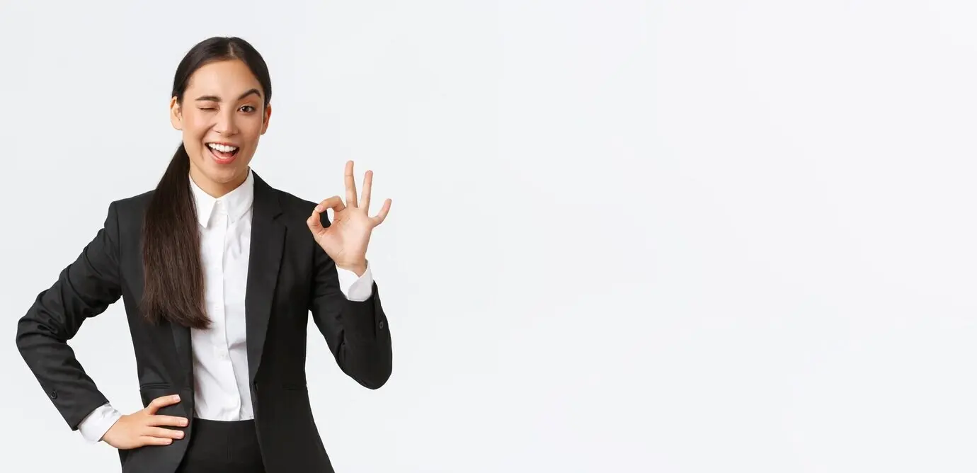 Durchsetzungsfähige, selbstbewusste junge asiatische Geschäftsfrau hat alles unter Kontrolle, zwinkert und zeigt das Okay-Zeichen, um zu ermutigen oder zu signalisieren, dass der Deal abgeschlossen ist, gute Arbeit geleistet wurde oder um Zustimmung auszudrücken; steht zufrieden.